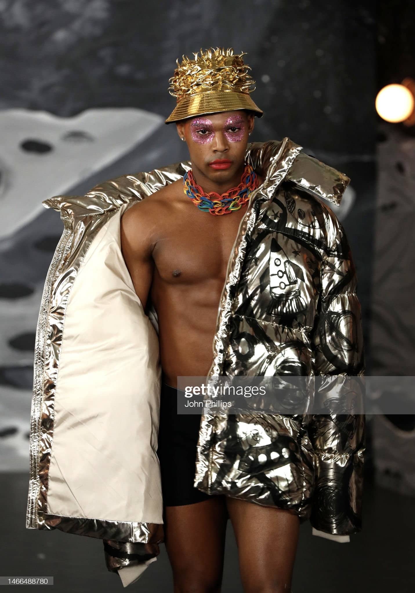 Man with shiny coat slightly open revealing chest, colorful necklace, and gold-colored hat